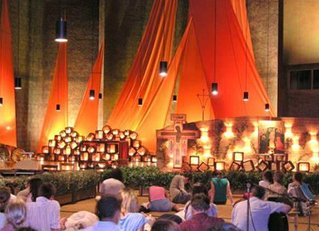 am Boden sitzende betende Menschen, Mann mit Gitarre, Kirche mit orangen Tüchern, viele Kerzen